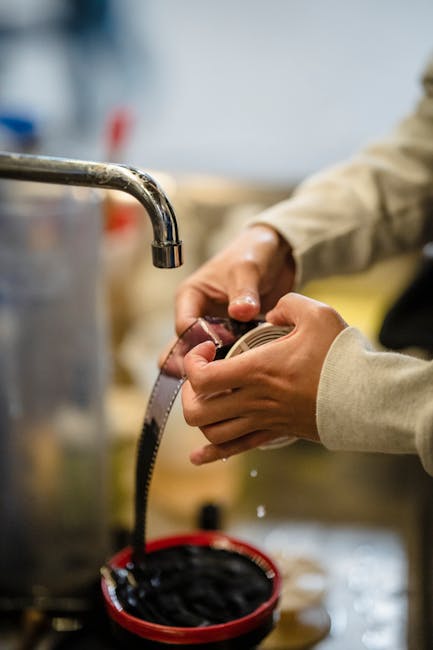 Close-up of a person’s hands wearing a beige long-sleeved shirt, holding a flexible measuring tape while inspecting or measuring a coiled black cable or flexible hose. The background shows a stainless steel sink or appliance with water running from a spout, suggesting a kitchen or utility area. The person is positioned to the right of the image, with the measuring tape extending towards the lower left. Nearby, there is a red container or bowl with additional black cables or hoses. The environment appears to be a workspace with various items and equipment related to home repairs or packing, fitting with the context of furniture transport and house removals. This scene may depict the checking or preparing of objects for a home relocation service by Movers Ilford, emphasizing careful handling during packing and moving processes.