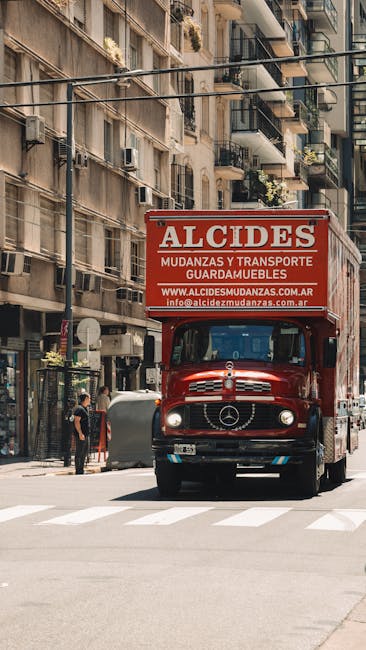 A large red moving truck with the branding 'ALCIDES Mudanzas y Transporte Guardamuebles' parked on a narrow urban street in Ilford, surrounded by multi-storey residential buildings with balconies and potted plants. The truck is positioned facing forward, with the Mercedes-Benz logo visible on the front grille. The vehicle is in the process of being loaded or unloaded, with a person standing nearby and items possibly being moved into or out of the truck. The street features a zebra crossing, with pedestrians and local residents visible in the background. The scene captures elements of home relocation logistics, including professional furniture transport and packing processes, relevant to house removals services provided by Movers Ilford. Bright daylight illuminates the scene, enhancing the clarity of the objects and environment.