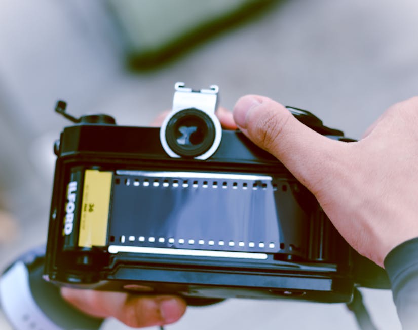 A close-up view of a person's hands holding a vintage camera with a black body and silver top, positioned outdoors on a cloudy day. The person’s left hand grips the camera, while their right hand adjusts the viewfinder or focus ring. The camera has a film compartment visible with a yellow Kodak film cartridge inserted, and the viewfinder lens is centered at the top. In the background, blurred elements suggest an outdoor environment, possibly a driveway or street, where moving or packing activities might occur. This scene illustrates careful handling during home relocation or moving preparations, with a focus on capturing moments during the packing or loading process, relevant to furniture transport and moving logistics. Movements or actions such as preparing to load packages or equipment related to house removals are implied by the image context, and the camera itself serves as a metaphor for documenting packing and moving procedures, connected to the services offered by Movers Ilford.