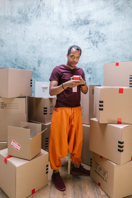 A man standing inside a room surrounded by stacked brown cardboard moving boxes of various sizes, some with red and black tape, and one partially open. He is wearing a maroon T-shirt and bright orange trousers, holding a smartphone and looking at the camera. The background features a textured light blue wall and a wooden floor, indicating a home or storage area used in the home relocation or furniture transport process, with the boxes prepared for packing and moving. This scene reflects the packing and loading stage of a house removals service, illustrating the careful organization required during professional furniture transport and moving logistics, as provided by Movers Ilford.