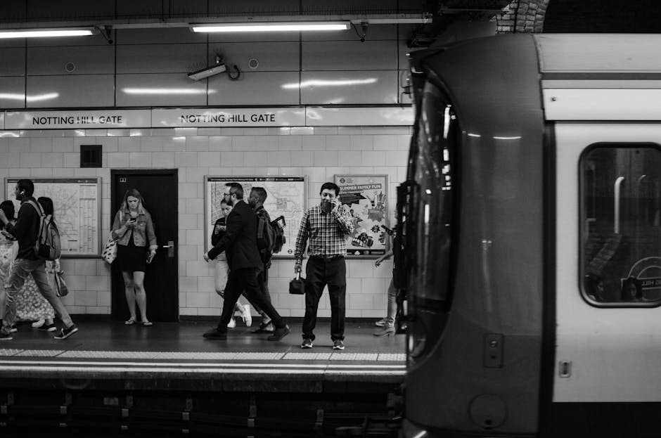 A black-and-white photograph of the Notting Hill Gate underground station platform, showing several passengers standing and walking near the tiled wall, which features signs and maps. The station's name, 'Notting Hill Gate,' is displayed on multiple signs above the passengers. To the right, part of a London Underground train is visible, with its doors closed. Passengers are dressed in casual and business attire, some carrying backpacks or shoulder bags, reflecting typical commuter activity. The platform has a safety line along the edge, and the area is well-lit with overhead lighting. This scene captures the daily flow of commuters, highlighting the typical environment where moving and packing logistics could be arranged by Movers Ilford, especially when coordinating home relocation or furniture transportation to or from the station.