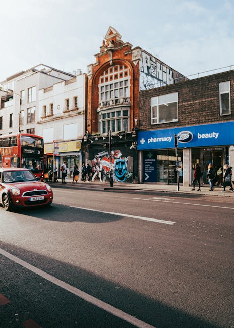 The image shows a busy street scene in Ilford with several pedestrians walking along the pavement and a red double-decker bus and a small red car driving on the road. The buildings facing the street include a prominent historic-looking structure with a decorative red brick façade, large arched window, and ornate architectural details at the top. Adjacent storefronts feature modern signage, including a pharmacy and a beauty shop, both with large glass windows displaying interior products. In front of the stores, people are entering or exiting, with some standing near the entrance. The scene is well-lit, indicating daytime with clear weather. This setting exemplifies typical urban high street activity, ideal for illustrating home relocation or furniture transport services provided by Movers Ilford during house moves or packing and loading processes, capturing the environment in which furniture transfer and moving logistics take place.