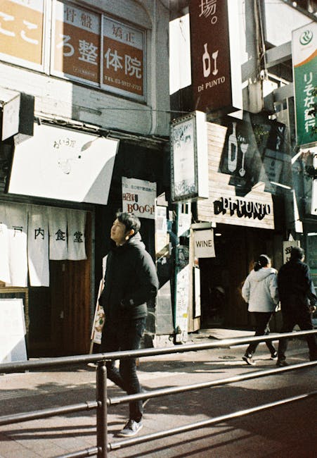 A busy urban street scene showing pedestrians walking past various Japanese restaurant signs and storefronts, with one man in dark clothing walking along the pavement near a metal railing. The buildings are multi-story with signage in Japanese characters and some in English, advertising dining establishments and wine shops. Inside the scene, there are cardboard boxes, plastic wrapping, and packing materials visible near a storefront entrance, indicating preparations for a home relocation or moving service. The natural daylight highlights the details of the storefronts and the movement of people, reflecting an active high street environment where furniture transport and packing are ongoing, consistent with the context of house removals in a busy city setting, as noted on the Movers Ilford website.