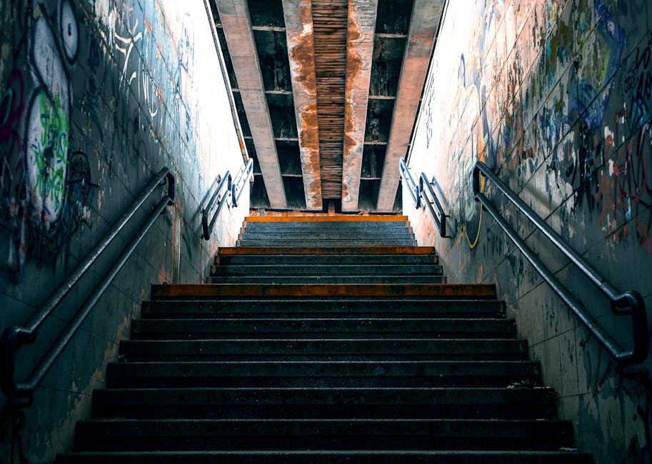 A set of underground staircase steps leading upwards, with black metal handrails on both sides. The walls adjacent to the stairs are covered in graffiti with vibrant colors, including green, blue, and red. The steps are made of dark material, possibly concrete or metal, with visible wear and some rust or dirt. At the top of the stairs, an industrial-style ceiling shows exposed wooden beams and metal supports. The lighting is dim, with natural light filtering from above, illuminating the upper part of the staircase. This scene depicts an urban environment, suitable for a home relocation or moving process involving stairs, and underscores the importance of furniture transport and packing during a house move, as handled by Movers Ilford.