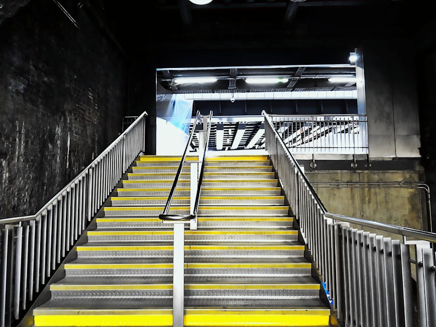 A wide view of an indoor staircase with metal handrails and yellow safety strips along each step, leading up to an open upper level in a modern building. The staircase is constructed from metal, with a removable middle handrail, indicative of support during moving or heavy lifting. The surrounding environment features dark, textured walls and exposed overhead pipework, typical of a commercial or parking facility. Natural light streams in from the upper open area, illuminating the stairs and highlighting the industrial design. This setting illustrates the kind of access challenges faced during home relocation, especially when navigating narrow routes or staircases, which professionals like Movers Ilford address with tailored loading and packing solutions, including the safe transport of furniture, boxes, and appliances. The overall scene emphasizes the importance of planning for difficult access points in residential moves within urban environments.