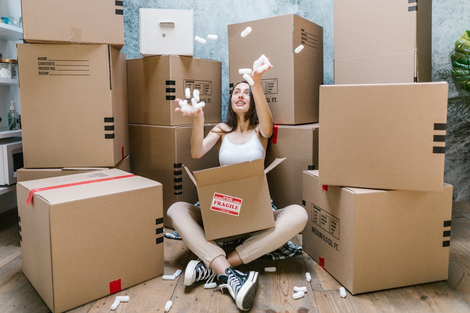 A young woman with long dark hair, sitting cross-legged on a wooden floor inside a room, is surrounded by large cardboard moving boxes, some with red tape and label stickers. She is wearing a white ta