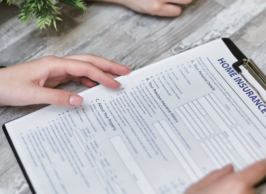 A close-up image of a person's hands holding a clipboard with a printed home insurance legal checklist, placed on a light wooden surface. The document contains headings such as 'A. Your Personal Detai
