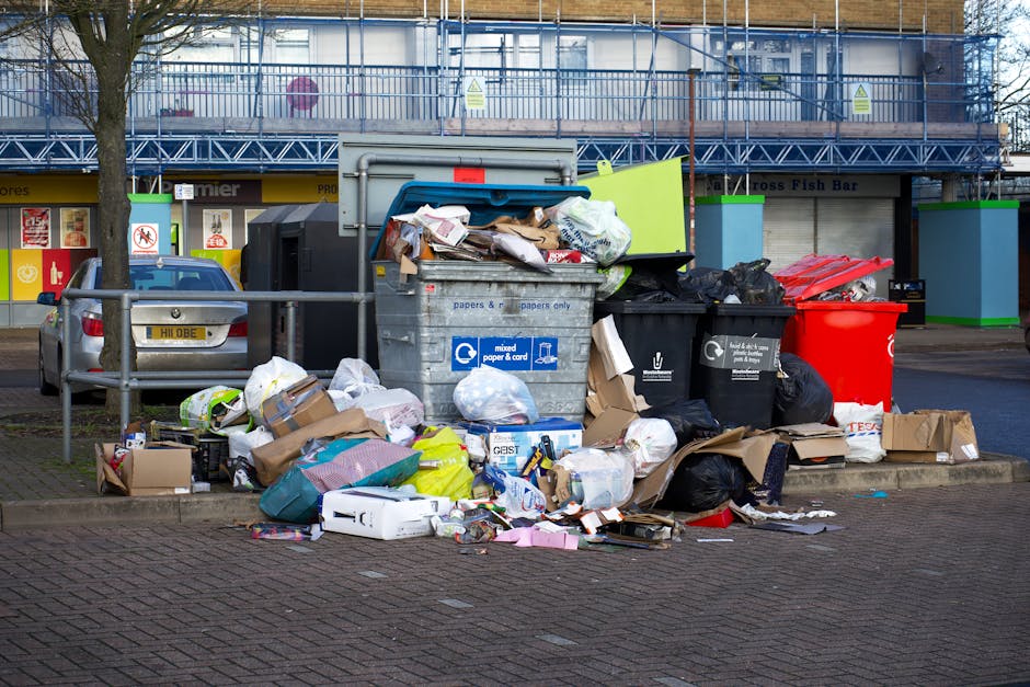 A waste collection operative wearing a high-visibility vest and dark clothing is operating a large, white waste disposal vehicle with red and orange striped reflective markings on the back. The vehicl