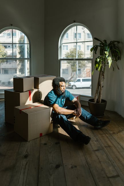 A man with curly hair and a beard, dressed in a blue t-shirt and dark jeans, sits on a wooden floor in a room with large arched windows that let in natural daylight. The windows reveal a view of nearby buildings and a street with parked cars outside. Next to him are several cardboard boxes, some sealed with red tape, indicating the packing process for a house removal. The boxes are stacked against a light-colored wall, and in the corner, there is a tall potted plant with broad green leaves. The room appears to be in the midst of a home relocation, with the man taking a brief rest during packing and moving activities. The scene highlights the typical environment of furniture transport and packing involved in house removals, with the natural lighting, interior setting, and packing materials all supporting the context of professional moving services like those offered by Movers Ilford.